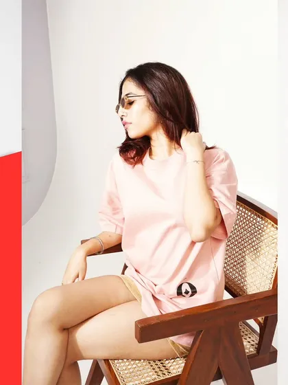 Posing and props are essential for showing apparel in a natural way. Here, the model's relaxed pose on a modern chair helps display the fit and drape of the oversized pink t-shirt, providing a lifestyle context that is perfect for product pages.