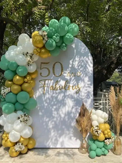 A '50 and Fabulous' birthday celebration in a garden, with a white arched backdrop and a green and gold balloon garland.