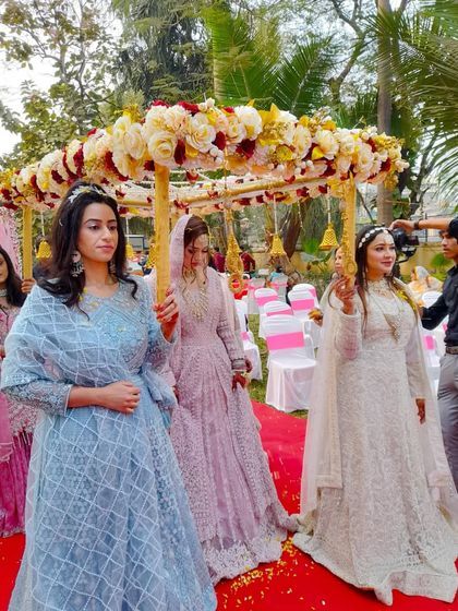 A bride makes her entrance under the yellow and maroon phoolon ki chadar, held by her bridesmaids. This shows how the canopy looks in action during an outdoor, daytime event.