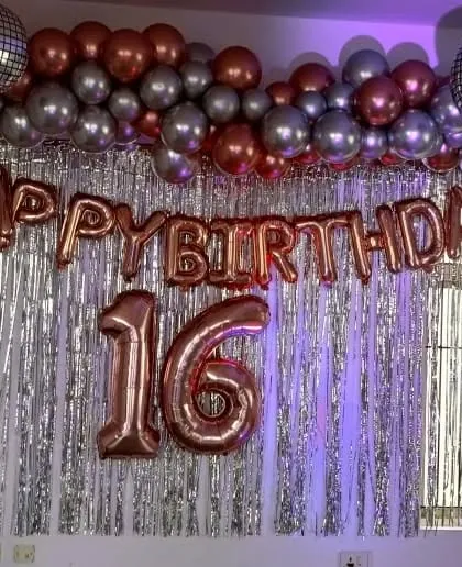 A 16th birthday decoration with a silver tinsel wall and rose gold foil balloons for the age and 'Happy Birthday' banner.