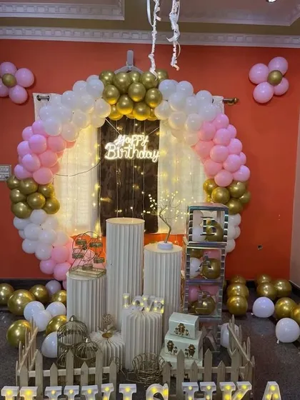 A sophisticated birthday setup with a white picket fence. This decoration features a pink, white, and gold balloon ring, pedestals for props, and illuminated letters spelling out the name 'HULSIKA'.