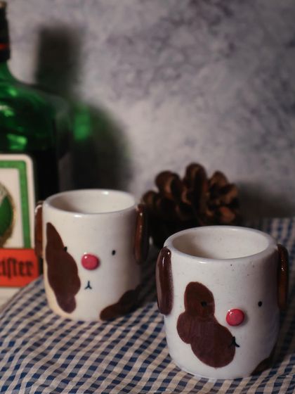 A pair of adorable dog-themed shot glasses from my 'Paw-try' collection, sitting on a checkered cloth. These small, handmade pieces are perfect for a little toast with friends.