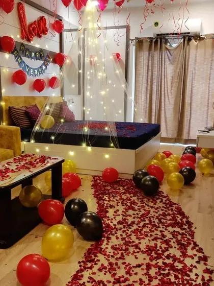 A complete romantic room transformation featuring a pathway of red rose petals. The decoration includes balloons in red, black, and gold, and a bed adorned with a fairy light canopy.