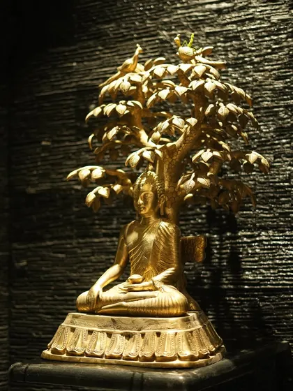 A detailed close-up of the golden Buddha statue, highlighting the intricate craftsmanship of the figure and the leaves of the tree above.