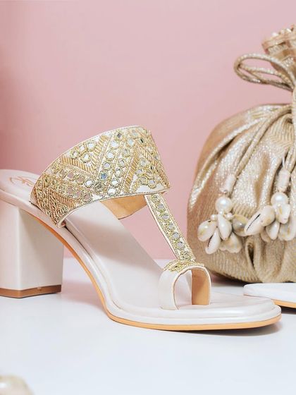 A beautifully styled shot of traditional Indian footwear paired with a golden potli bag. The soft pink background and elegant props create a festive and luxurious feel, perfect for occasion wear.