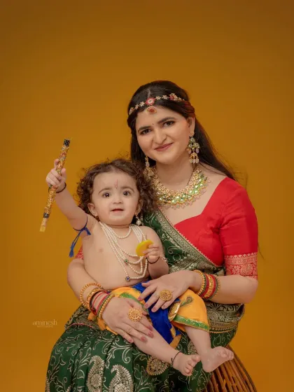 A lively shot of a baby Krishna raising his flute while being held by his mother. This image captures a moment of joy and celebration during the themed photoshoot.