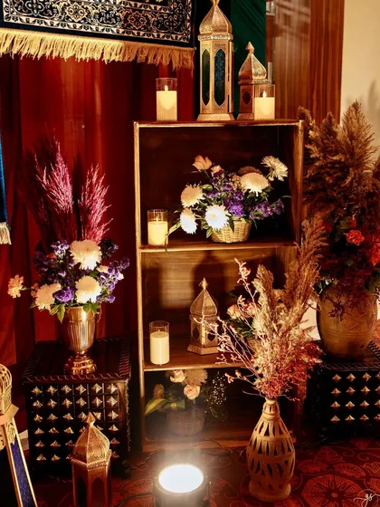 A cozy decor corner with a wooden shelf holding candles and flowers, set against a backdrop of rich red drapes and hanging rugs.