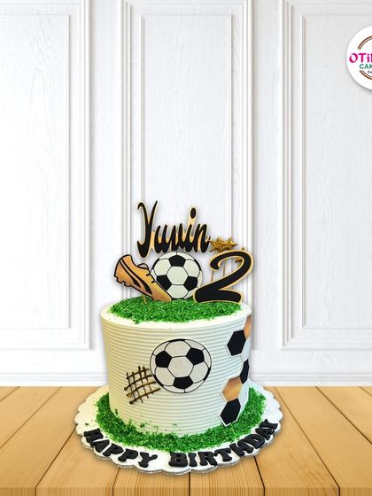 This football-themed cake is ready for kickoff. We decorated it with edible soccer balls, a golden shoe topper, and a grassy buttercream field, personalized for the birthday boy.