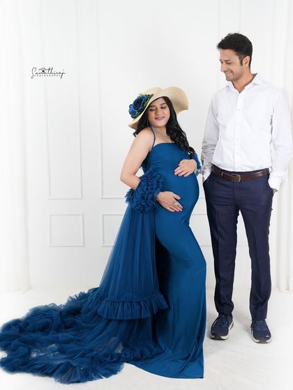 A stylish couple portrait with the mother-to-be in a blue gown and hat. The clean, modern studio setting is perfect for a chic and fashionable maternity shoot.