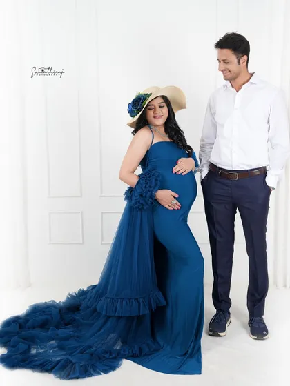 A stylish couple portrait with the mother-to-be in a blue gown and hat. The clean, modern studio setting is perfect for a chic and fashionable maternity shoot.