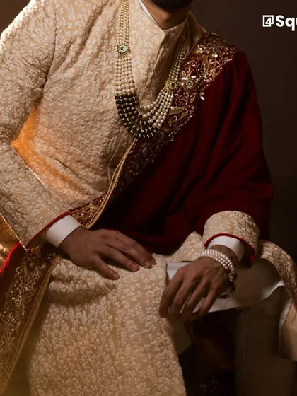 We craft elegant sherwanis that are custom-fitted to make you look and feel exceptional on your special day. This textured beige sherwani, paired with a deep red velvet shawl and pearl accessories, exemplifies the regal aesthetic we deliver for discerning grooms.