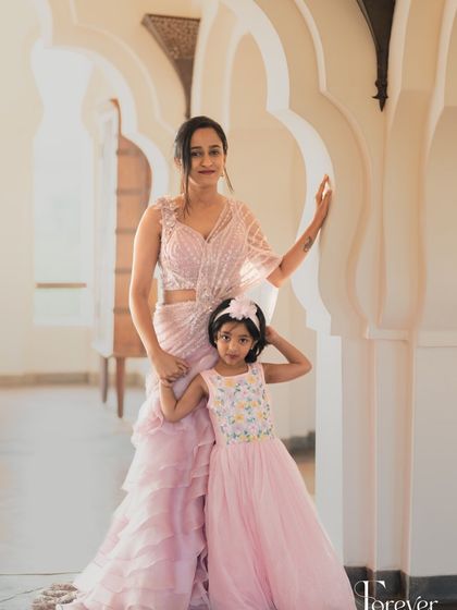 A mother and daughter in matching pastel pink outfits. The mother wears a modern ruffled saree, while her daughter is in an adorable matching gown, showing how I can create coordinated looks for all ages.