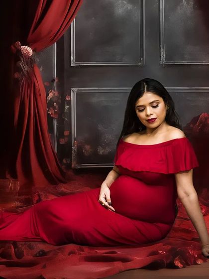 A serene, seated portrait from a fine art maternity session. The setup includes a floor covered in red fabric and petals, with dramatic curtains and florals in the background, creating a rich and romantic atmosphere.