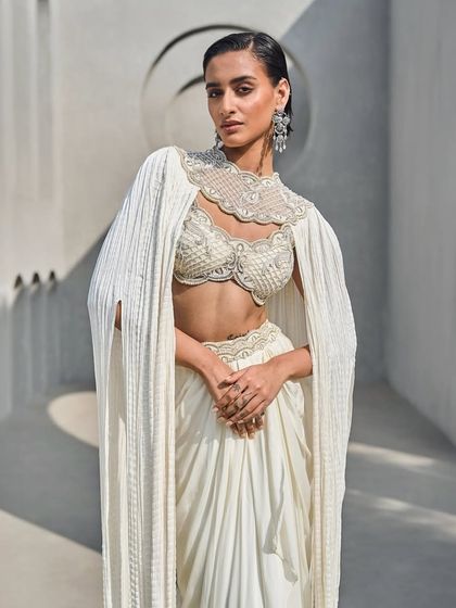 Ivory in motion, where sculpted glamour meets fluid grace. This ensemble features crushed cape sleeves, an embellished bustier, and a cinched drape.