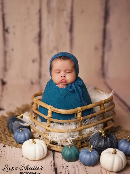 A baby boy swaddled snugly in a deep teal wrap, wearing a matching bonnet and sleeping in a wicker basket.
