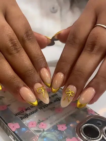 An elegant yet playful design with a gold chrome French tip and 3D gold bow accents on almond-shaped nails.