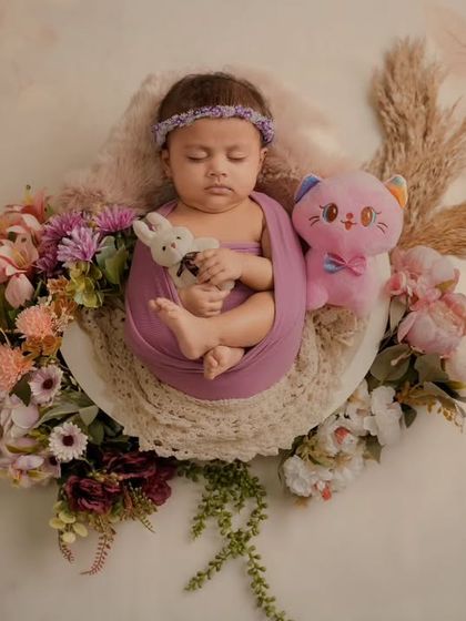 A sleeping newborn is nestled in a floral arrangement with a pink teddy bear, set against a soft, floral-patterned backdrop.