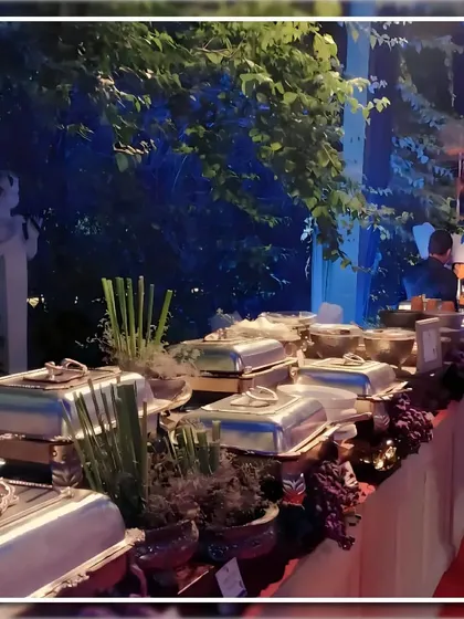 A buffet table set for an evening event in a garden. The silver chafing dishes are surrounded by lush foliage and subtle lighting, creating an enchanting dining experience.