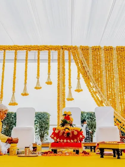 A traditional Haldi ceremony in progress, with the priest seated before a stunning backdrop of marigold garlands. The setup is clean, vibrant, and respectful of tradition.