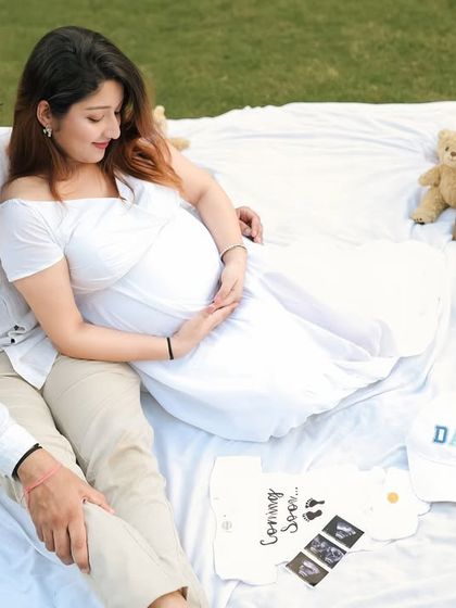An expecting couple enjoys a quiet moment during an outdoor picnic maternity shoot. Props like 'Mom' and 'Dad' hats, a 'Coming Soon' onesie, and ultrasound pictures add a personal touch.