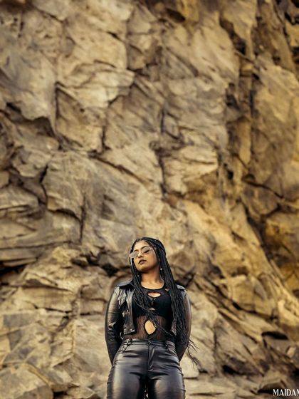 A wide shot of the model standing confidently against a massive rock wall. The scale of the background emphasizes her strong and commanding presence.