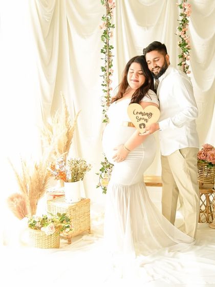 A sweet moment with the couple holding a "Coming Soon" sign. The all-white theme with soft floral and pampas grass accents creates a dreamy and romantic atmosphere.