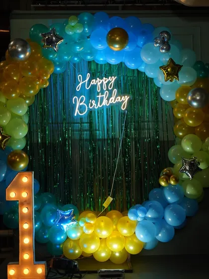A stylish first birthday decoration featuring a circular balloon arch, a lit-up number '1', and a neon 'Happy Birthday' sign. We take detailed shots of your decor.