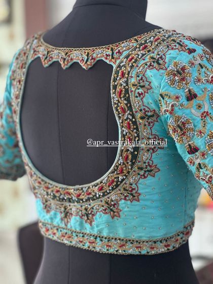 A heavily embroidered blouse in a striking sky blue, shown on a mannequin to highlight the dramatic back cutout. The entire surface is covered in floral zardosi work.