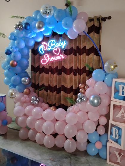 A gender reveal or baby shower setup with a half-moon balloon arch in pink and blue. The 'BABY' blocks and 'Baby Shower' neon sign make it a great photo spot.