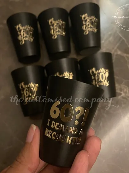 Cheers to 60 years! These custom black shot glasses are perfect for a milestone birthday party, featuring cheeky phrases like "60?! I DEMAND A RECOUNT!" in elegant gold lettering.