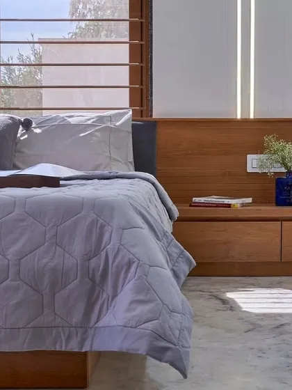 A calm and restful bedroom corner. The low wooden bed frame extends into a convenient bedside bench, and the large windows with slatted blinds allow for soft, diffused sunlight.
