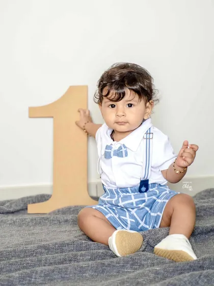 A classic first birthday portrait. He sits confidently next to the number '1', dressed in a smart outfit with suspenders and a bow tie.