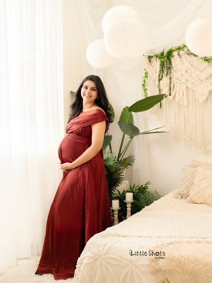 A full-length portrait of an expecting mother in a deep red dress. She stands in a bright, boho-inspired room with large plants and natural light, smiling gently.