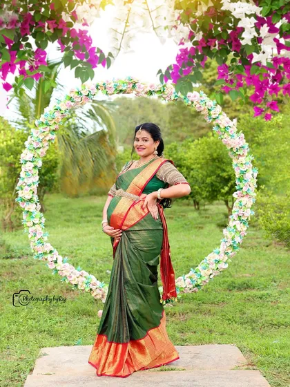 A stunning outdoor portrait of a mother-to-be in a green and red silk saree, framed by a circular floral prop.