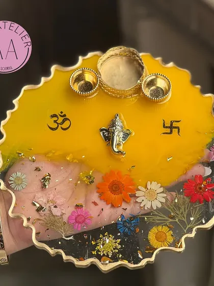 This pooja thali features a bright yellow top, symbolizing happiness and auspiciousness. The clear bottom half is filled with colorful preserved flowers and gold flakes.