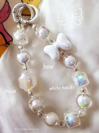 An angelic white beaded bag charm with iridescent beads that catch the light beautifully. It includes a white bow and a paw print charm for a touch of sweetness.