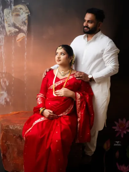 A stunning fine art maternity portrait. The mother-to-be, dressed in a vibrant red saree, is the picture of grace against a backdrop reminiscent of a classical painting.