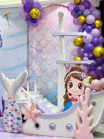 A detailed shot of the mermaid-themed party decor, featuring a boat prop with a mermaid character peeking out, surrounded by purple and gold balloons.