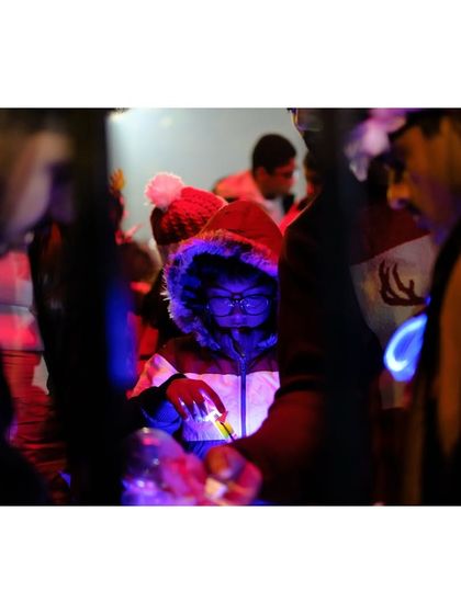 A child in a hooded jacket looks on, their face illuminated by the blue and red lights of Christmas decorations. This candid shot captures the wonder and magic of the festival through a child's eyes.