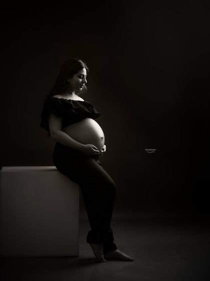 A quiet and contemplative low-key portrait. The client is seated, and the soft light gently falls on her and her bare bump, creating a peaceful and artistic black and white image.