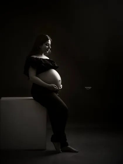 A quiet and contemplative low-key portrait. The client is seated, and the soft light gently falls on her and her bare bump, creating a peaceful and artistic black and white image.