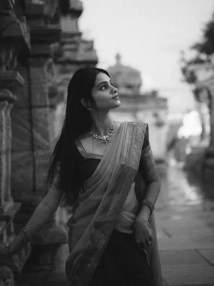 A black and white portrait of a model in a saree, taken at a temple. The monochrome treatment emphasizes the textures, shadows, and timeless elegance of the scene.