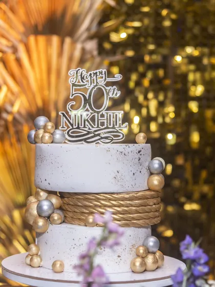 A close-up of the elegant two-tier cake for Nikhil's 50th birthday. The design features a textured white finish, a gold rope detail, and scattered gold and silver spheres.