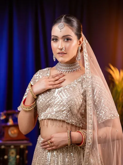 A graceful and polished look featuring a model in a shimmering silver-white lehenga. The soft studio lighting highlights the subtle glow of the makeup and the intricate details of the diamond necklace and traditional bangles, creating a perfect sangeet or reception look.