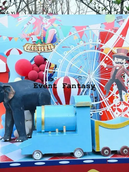 A different view of Neel's carnival party, showing a blue train prop in front of a backdrop filled with circus animals and a big top tent.