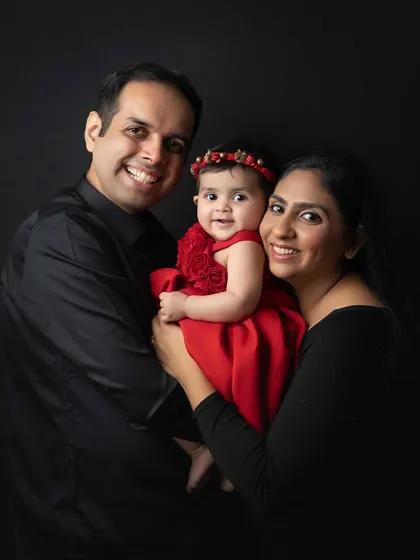 A classic portrait of a smiling family. The parents hold their daughter lovingly between them, creating a timeless image against a dramatic black background.
