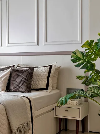 A touch of green from a large Monstera plant brings life to this serene, neutral bedroom corner. The paneled wall and quilted bedding add layers of subtle texture.