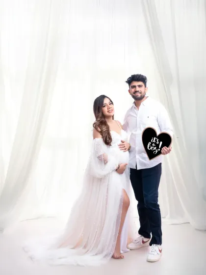 It's a boy! The happy couple makes their announcement with a heart-shaped sign, both dressed in white for a bright and celebratory portrait.