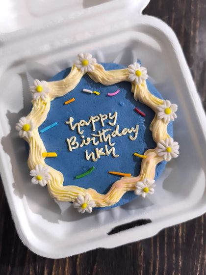 A trendy design featuring a wavy buttercream border. This blue bento cake is decorated with tiny, delicate fondant daisies, adding a touch of flair to a simple birthday message.