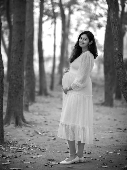 A stunning full-length solo portrait in black and white. The contrast between the white dress and the dark tree trunks creates a striking and artistic composition.